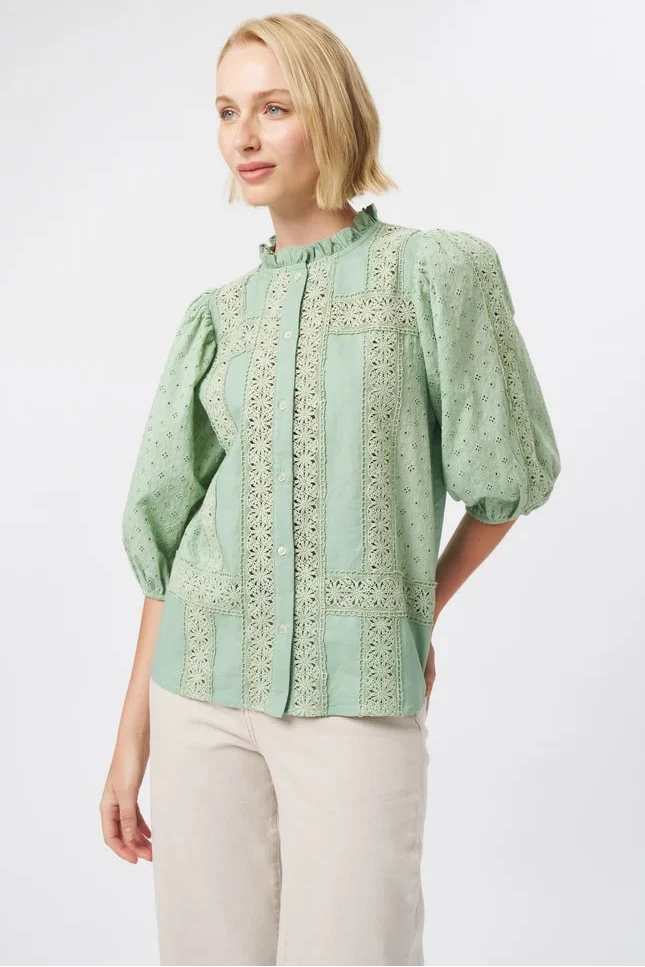 A woman modeling a sage green blouse with intricate floral embroidery and puff sleeves, paired with light beige trousers, against a plain light gray background.