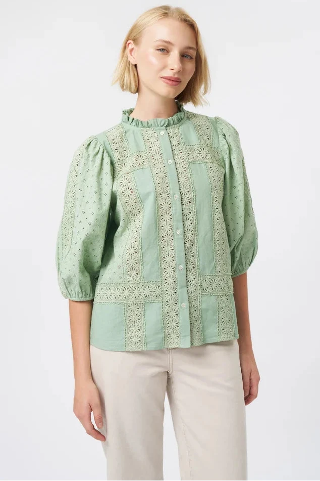 A woman modeling a light sage green blouse with intricate embroidery and puff sleeves, paired with beige trousers, against a plain white studio background.