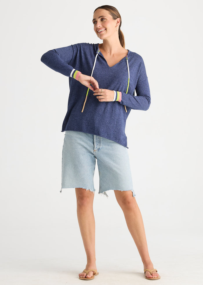 A woman modeling a navy blue heathered long-sleeve hoodie top with white drawstrings and colorful striped cuffs, paired with light-wash denim shorts and sandals, against a plain white studio background.