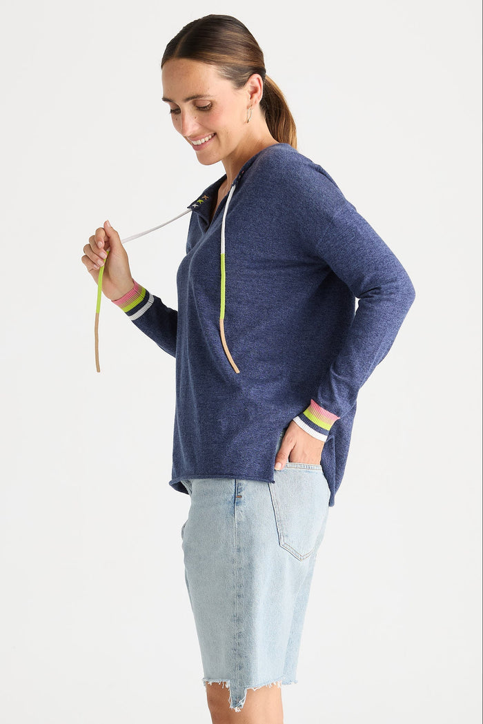 A woman modeling a heathered navy blue long-sleeve hoodie with white drawstrings and green and yellow striped cuffs, paired with a light denim skirt, against a plain white studio background.