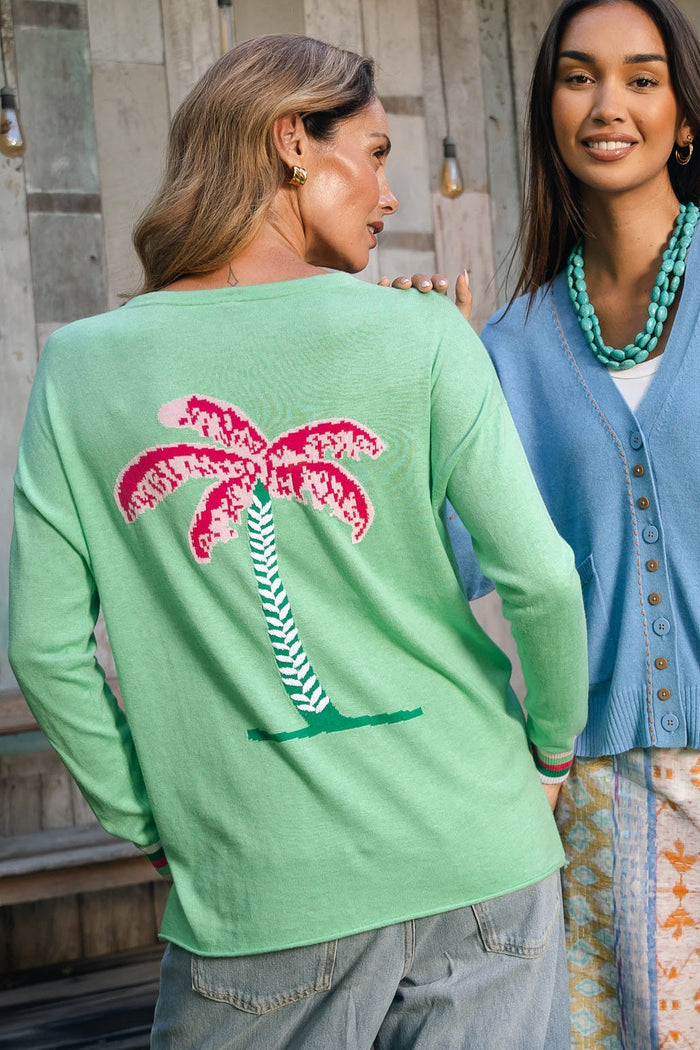 Two women modeling knit cardigans outdoors in front of a rustic wooden wall. The woman on the left wears a mint green cardigan with a large pink and white palm tree design on the back, paired with light blue jeans.