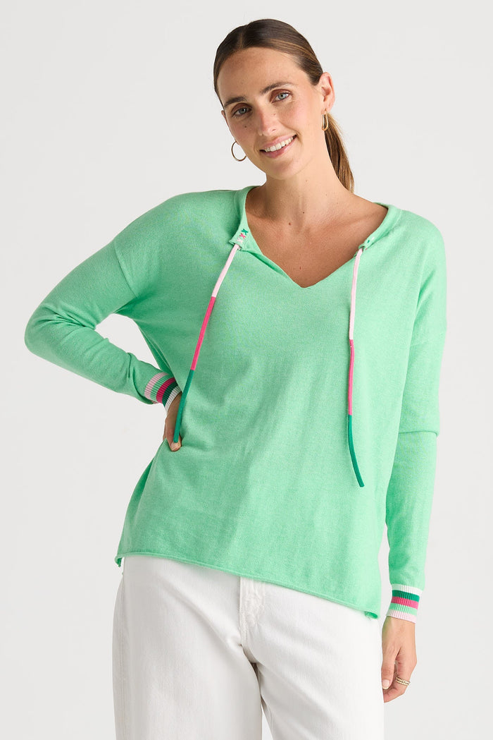 A woman modeling a mint green long-sleeve V-neck knit top with a drawstring hood and colorful striped cuffs, paired with white pants against a light gray studio background.