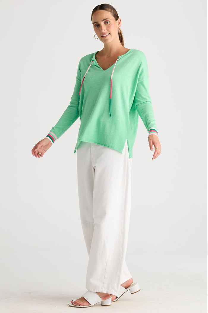 A woman modeling a mint green long-sleeve knit top with a drawstring hood, paired with white wide-leg pants and white slide sandals, against a plain white studio background.