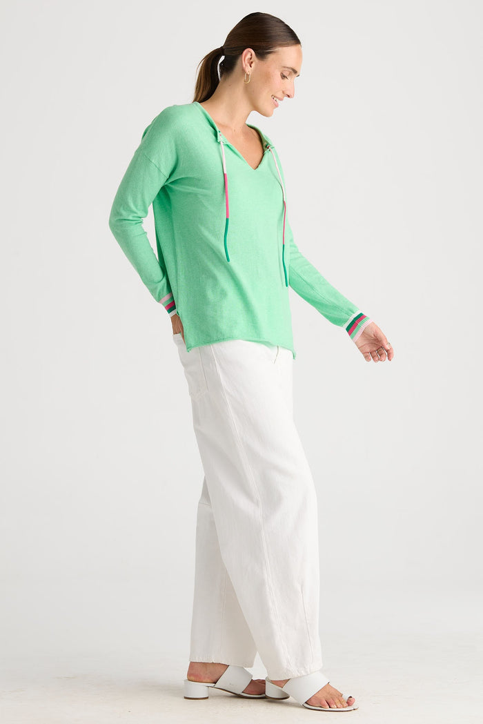 A woman modeling a mint green long-sleeve V-neck top with thin red and blue stripes along the armholes and cuffs, paired with white wide-leg pants and white sandals, against a plain white studio background.
