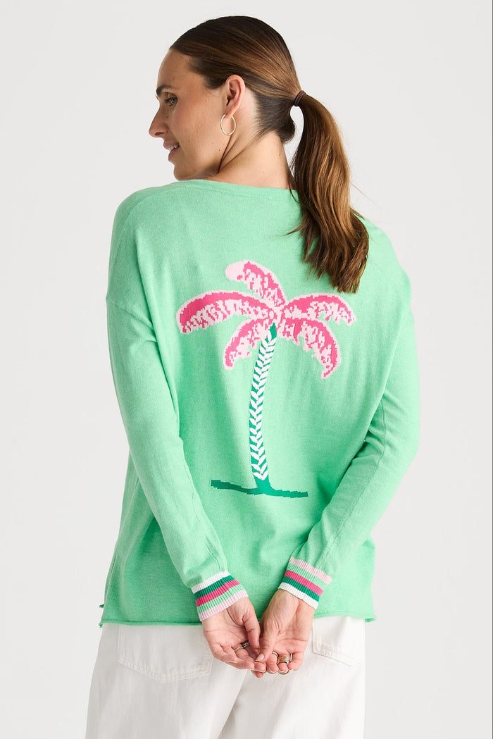 A woman shown from the back wearing a mint green knit sweater with a large pink palm tree graphic on the back and striped knit cuffs, paired with white pants against a light gray studio background.