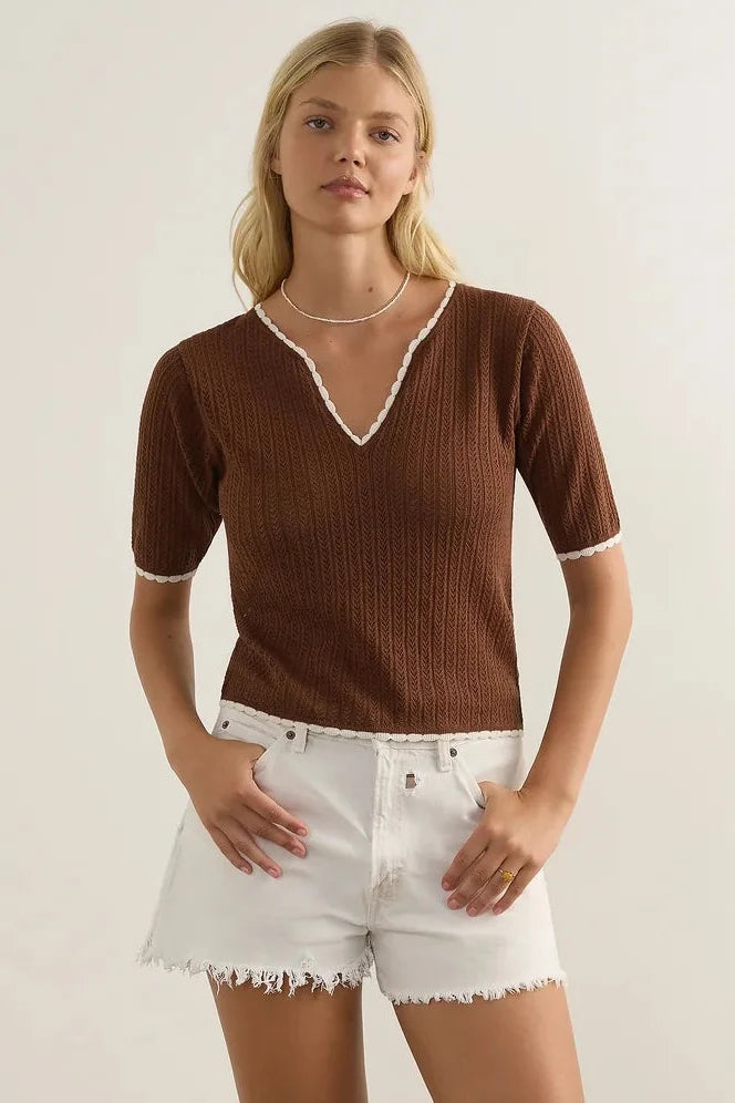 A woman modeling a brown ribbed short-sleeve V-neck top with white trim, paired with white frayed denim shorts and brown strappy sandals, standing against a plain off-white studio wall.