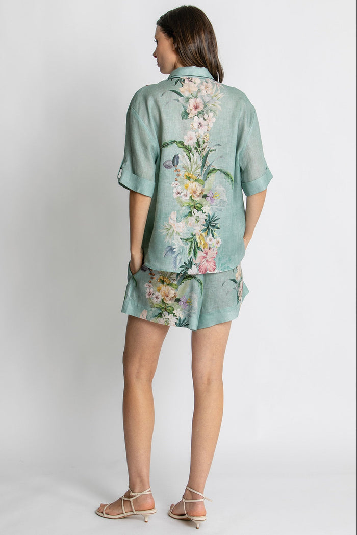 A woman shown from the back wearing a mint green short-sleeve button-up shirt and matching shorts set with a large multicolor floral print, paired with beige strappy sandals, against a light gray studio background.