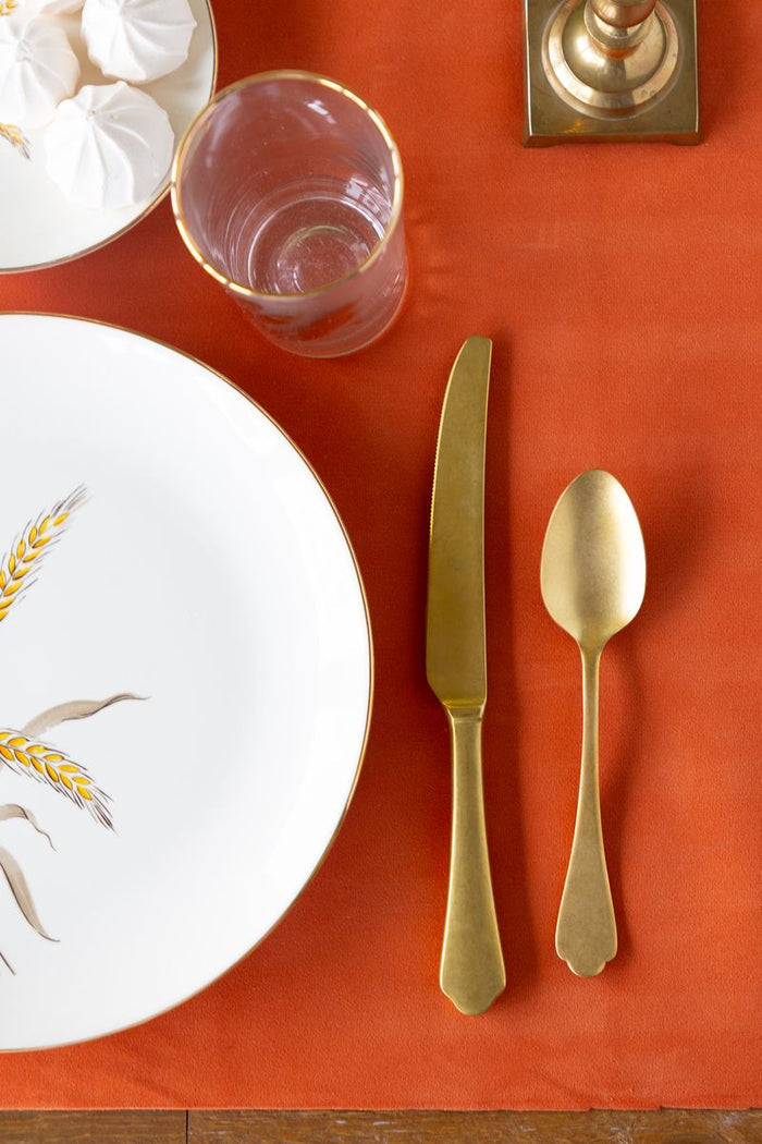 Table setting with white plate, gold cutlery, and napkin on an orange felted table runner.