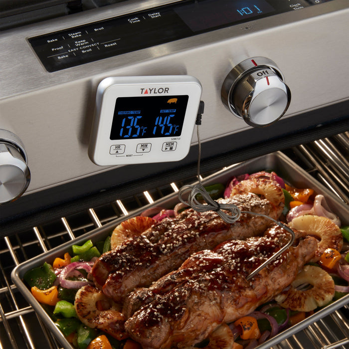 Taylor digital wired thermometer beside a grill with seasoned steak and vegetables cooking