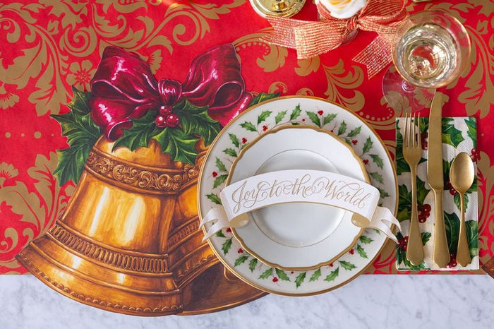 Christmas-themed place setting with gold bell, red bow, and 'Joy to the World' card on a red and gold patterned table runner.