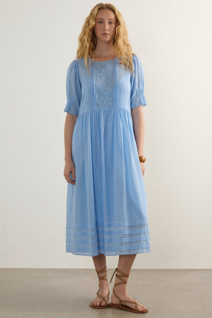 A woman modeling a light blue midi dress with short puff sleeves, a square neckline, and embroidered detailing on the bodice and hem, standing against a plain off-white studio background.