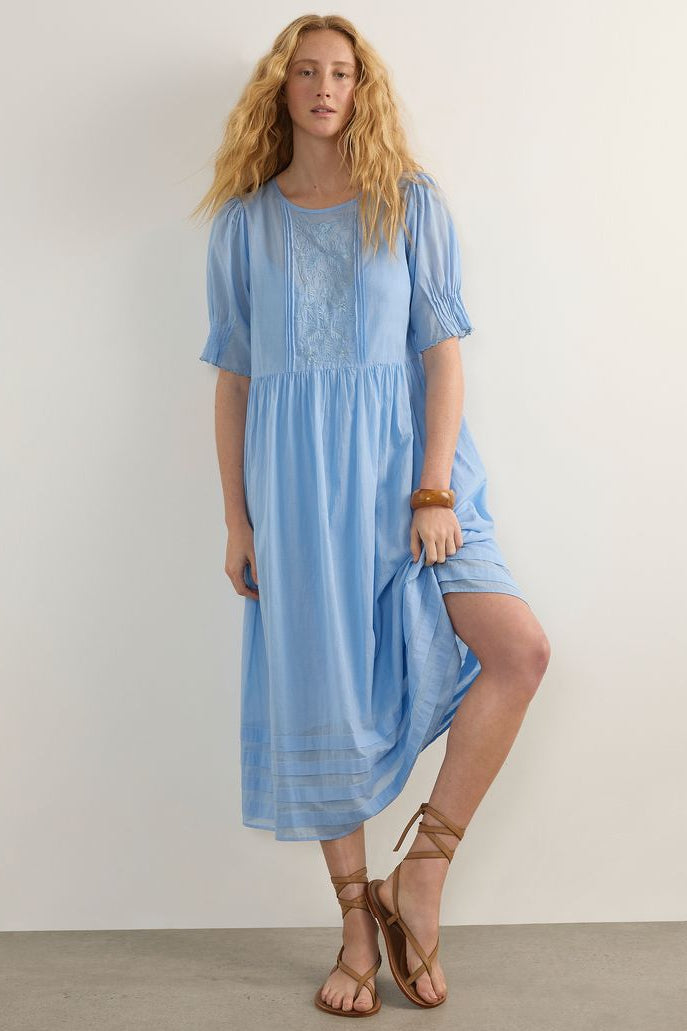 A woman modeling a light blue midi dress with short puff sleeves, a round neckline, and embroidered detailing on the chest, paired with brown strappy sandals against a plain off-white wall.