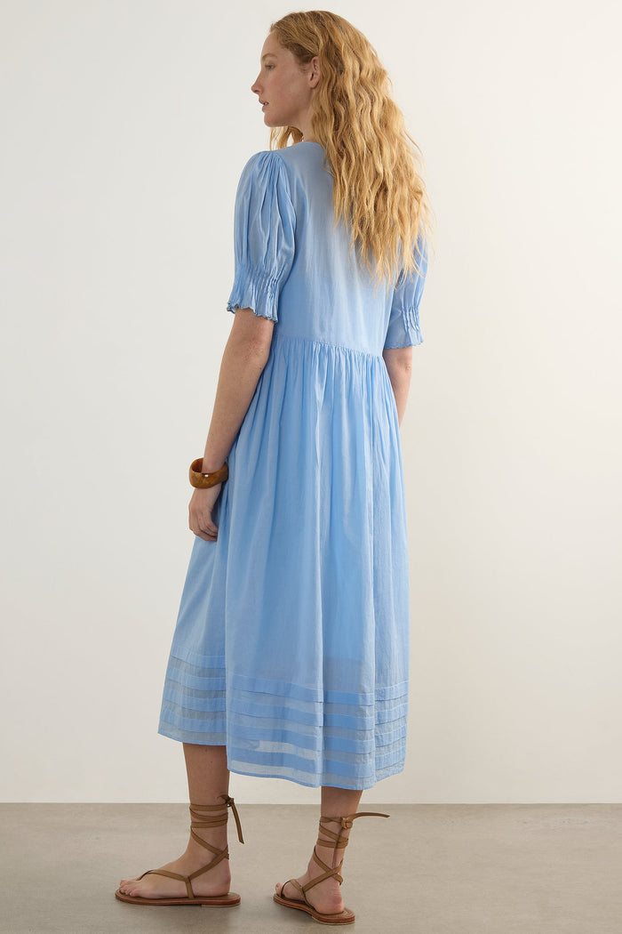 A woman with long wavy blonde hair is shown from the back wearing a light blue midi dress with puff sleeves and lace-up sandals, standing against a plain off-white wall.