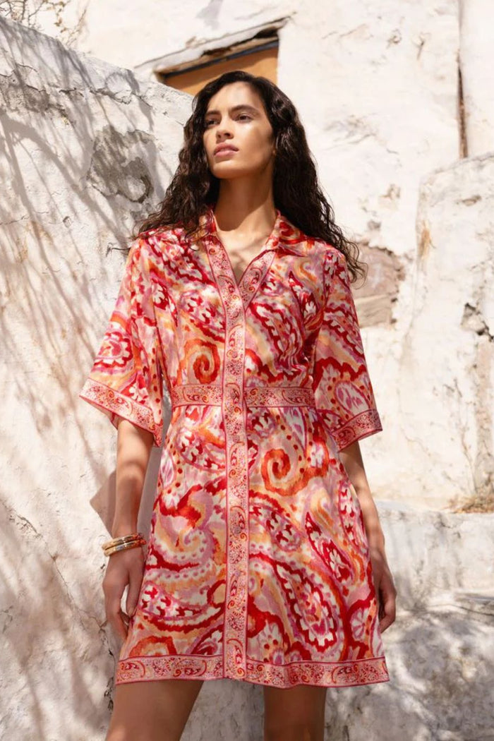 Woman wearing a red and orange patterned dress standing against a textured stone wall.