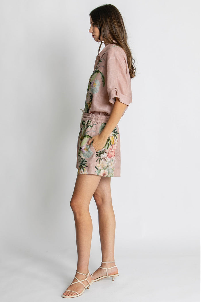 A woman modeling a dusty pink short-sleeve top and matching floral-print shorts against a light gray studio background.