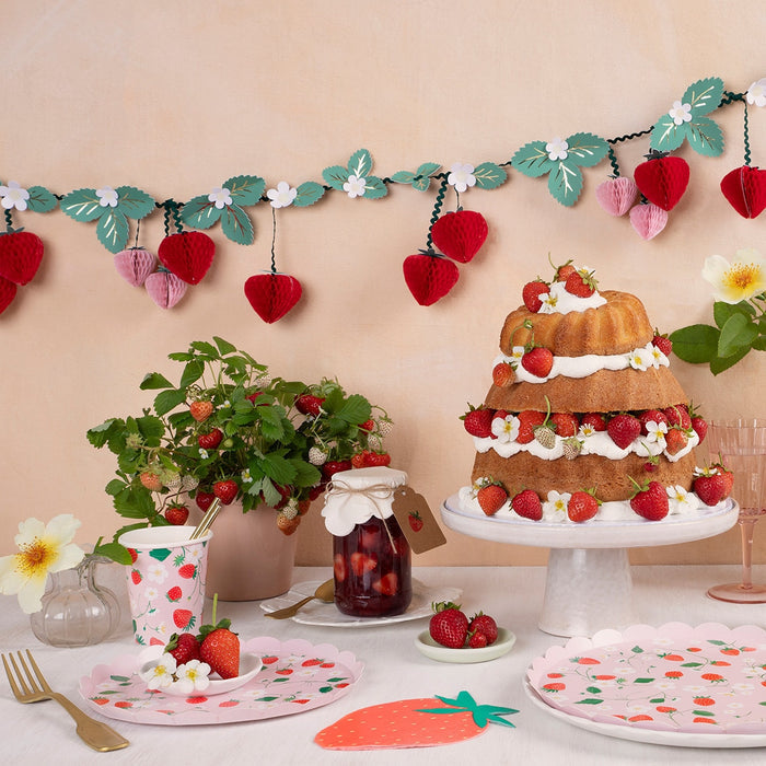 A decorative strawberry-themed table setting with a strawberry-patterned paper plates and cups as well as a strawberry banner, cake, and plant.