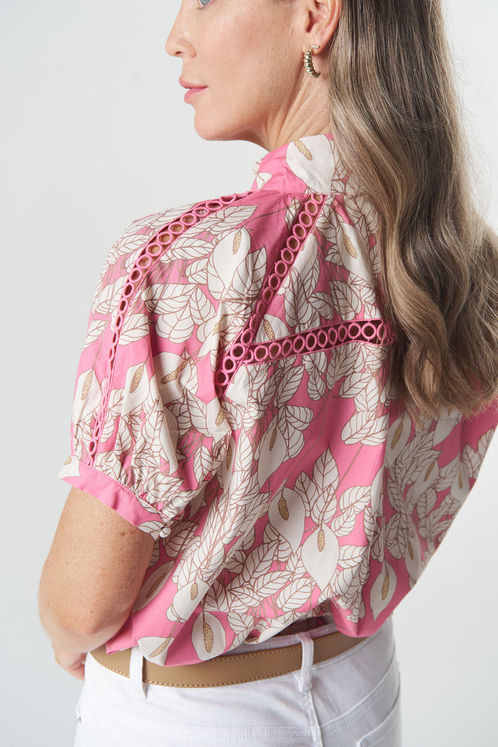 A woman shown from the back wearing a pink and beige floral print blouse with eyelet cutout details along the shoulders, paired with white pants.