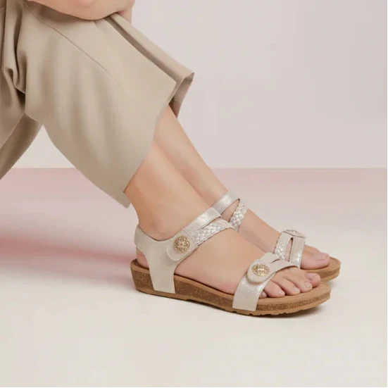 Woman sitting on a white surface wearing beige pants and sandals against a light pink background