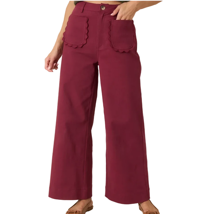 A person wearing high-waisted wide-leg pants in a deep burgundy or maroon color, featuring scalloped edges on the front patch pockets and a button closure, paired with tan flat shoes against a plain beige studio background.