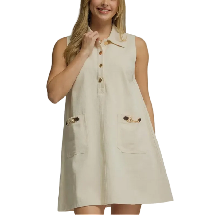 A woman modeling a sleeveless cream-colored linen dress with a button-down front, collar, and two front patch pockets with decorative brown and gold button details, set against a solid gray studio background.