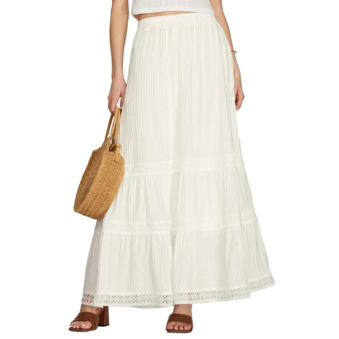 Woman wearing a white top and long white skirt holding a woven bag on a plain background