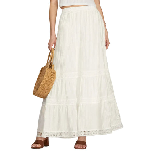 Woman wearing a white top and long white skirt holding a woven bag on a plain background