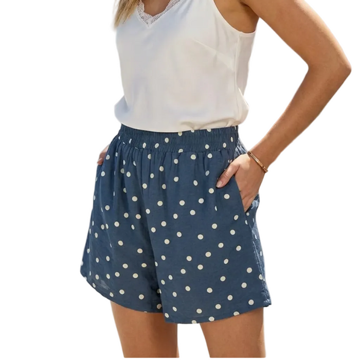 Woman wearing a white top and blue polka dot shorts standing on a sunlit terrace with ocean view.
